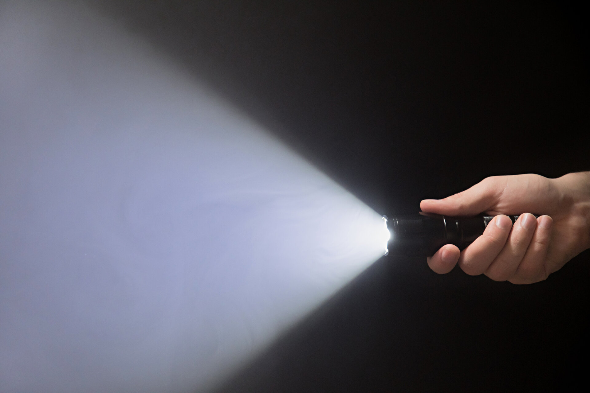 Black flashlight with a beam of light in male’s hand isolated from right side of the frame on black background copyspace